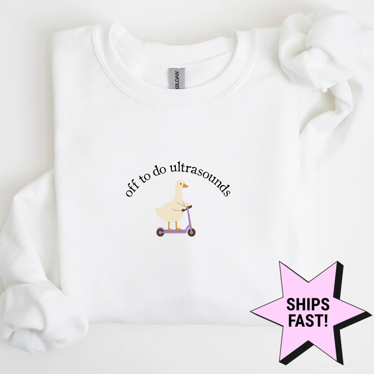White crewneck sweatshirt laid flat with a bunched sleeve, a goose on a purple scooter under arched off to do ultrasounds