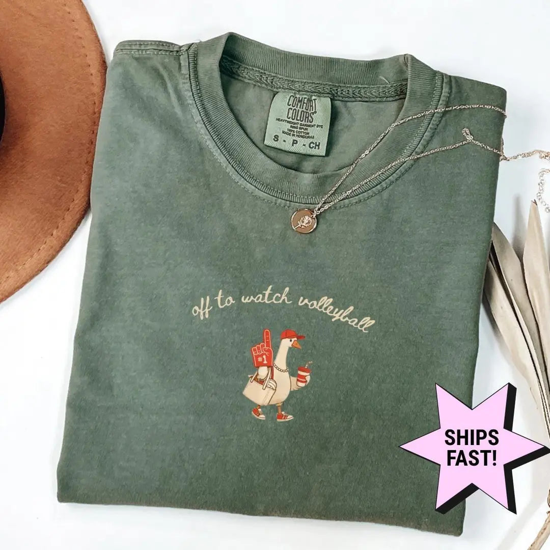 Folded Moss green Comfort Colors tee with the goose in a red cap and cursive volleyball text, laid flat with dried leaves
