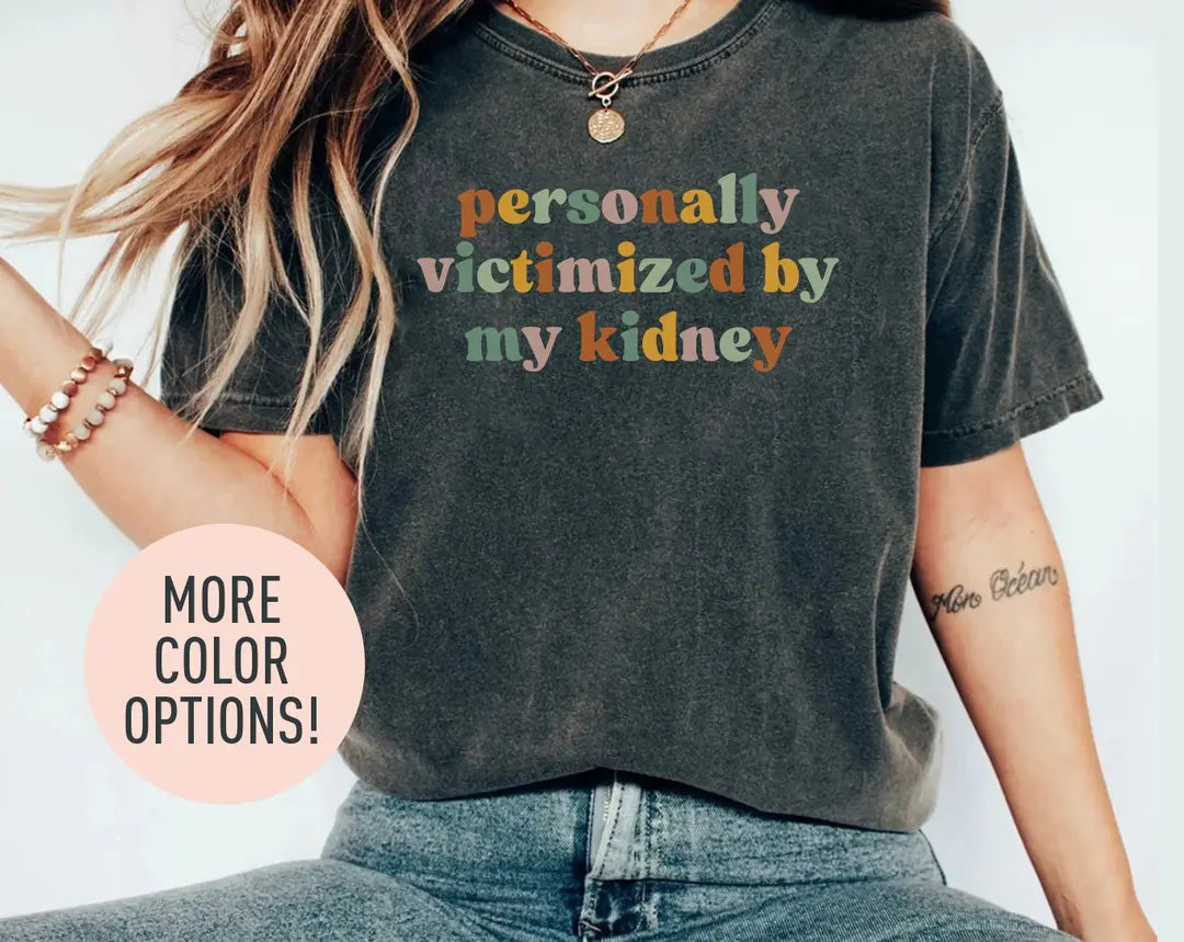 Woman in a dark gray Comfort Colors tee and jeans, bracelets, with personally victimized by my kidney in retro type