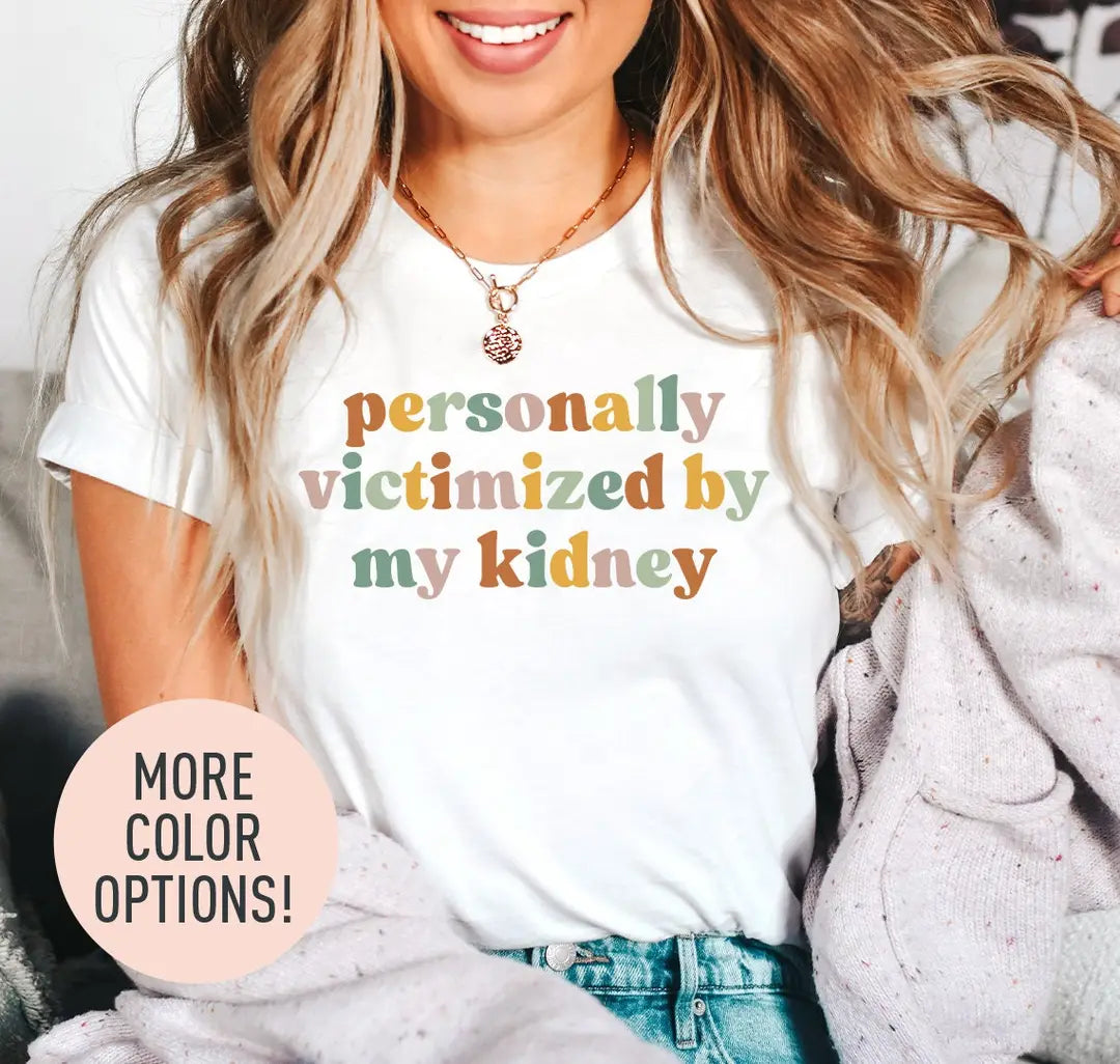 Woman in a white tee and a lilac cardigan, personally victimized by my kidney stacked in mustard, sage and rose letters