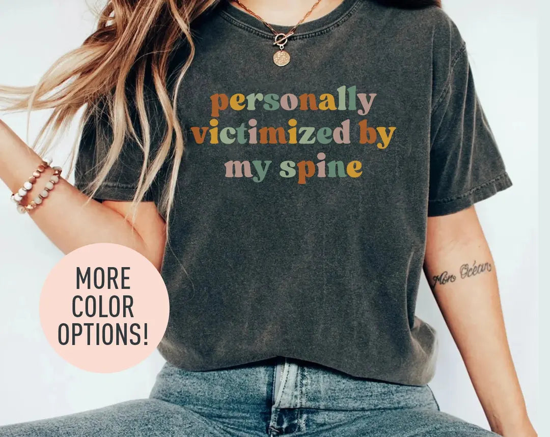Woman in a dark gray Comfort Colors tee and jeans, bracelets, with personally victimized by my spine in retro type