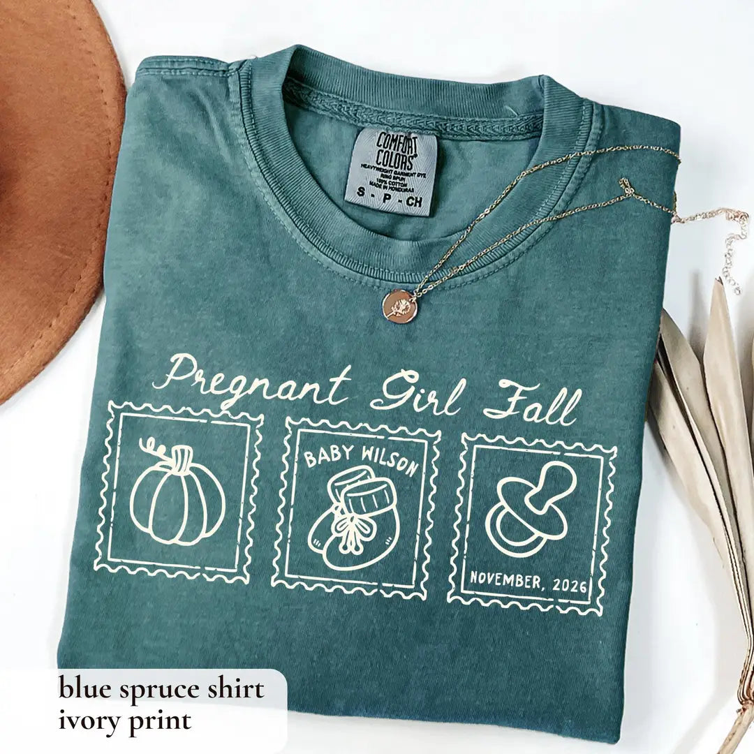 Folded Blue Spruce Comfort Colors tee with ivory pregnant girl fall script above a pumpkin, baby bootie and pacifier stamps