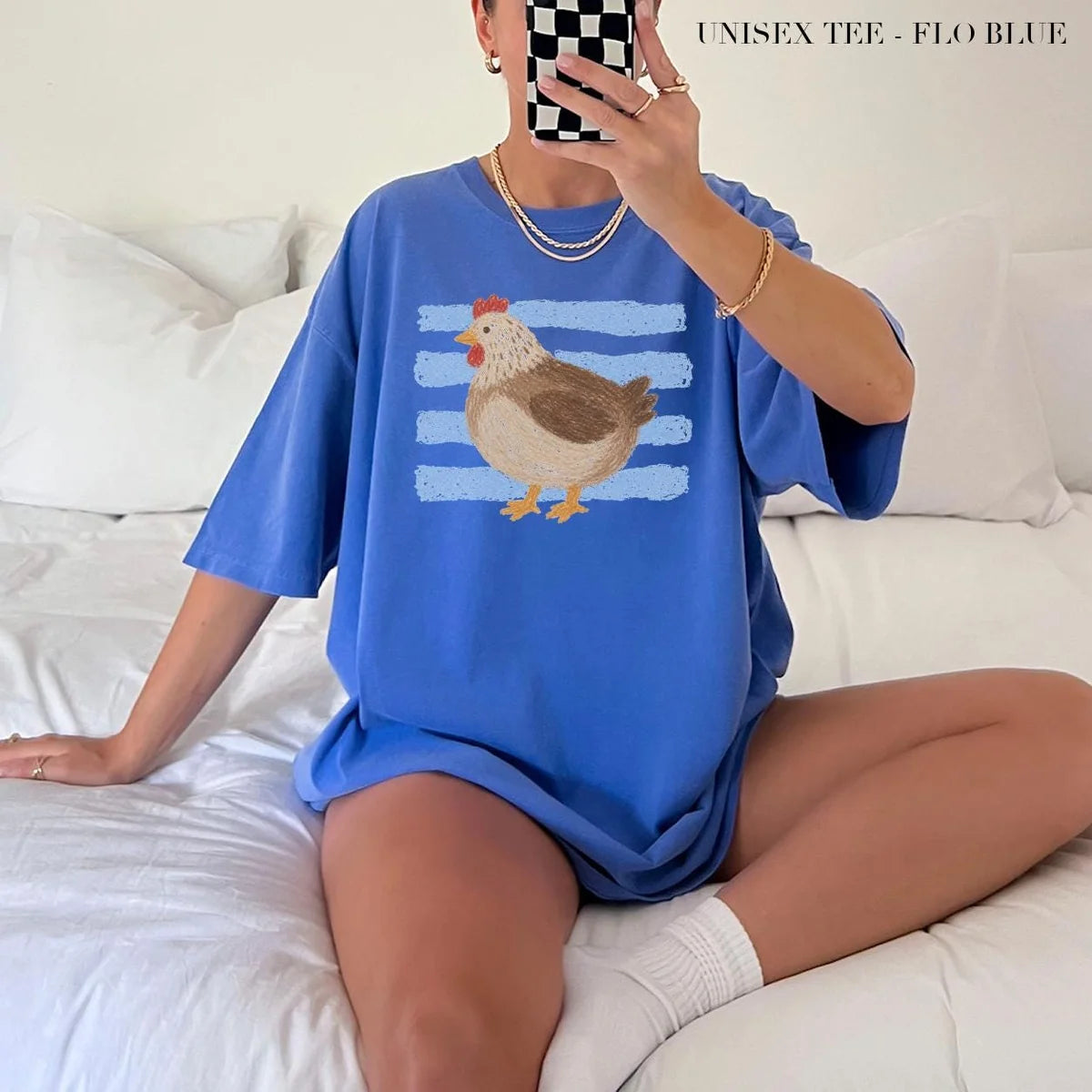 Woman in a Flo Blue Comfort Colors tee taking a mirror selfie on a bed, a small brown hen on blue stripes on the chest