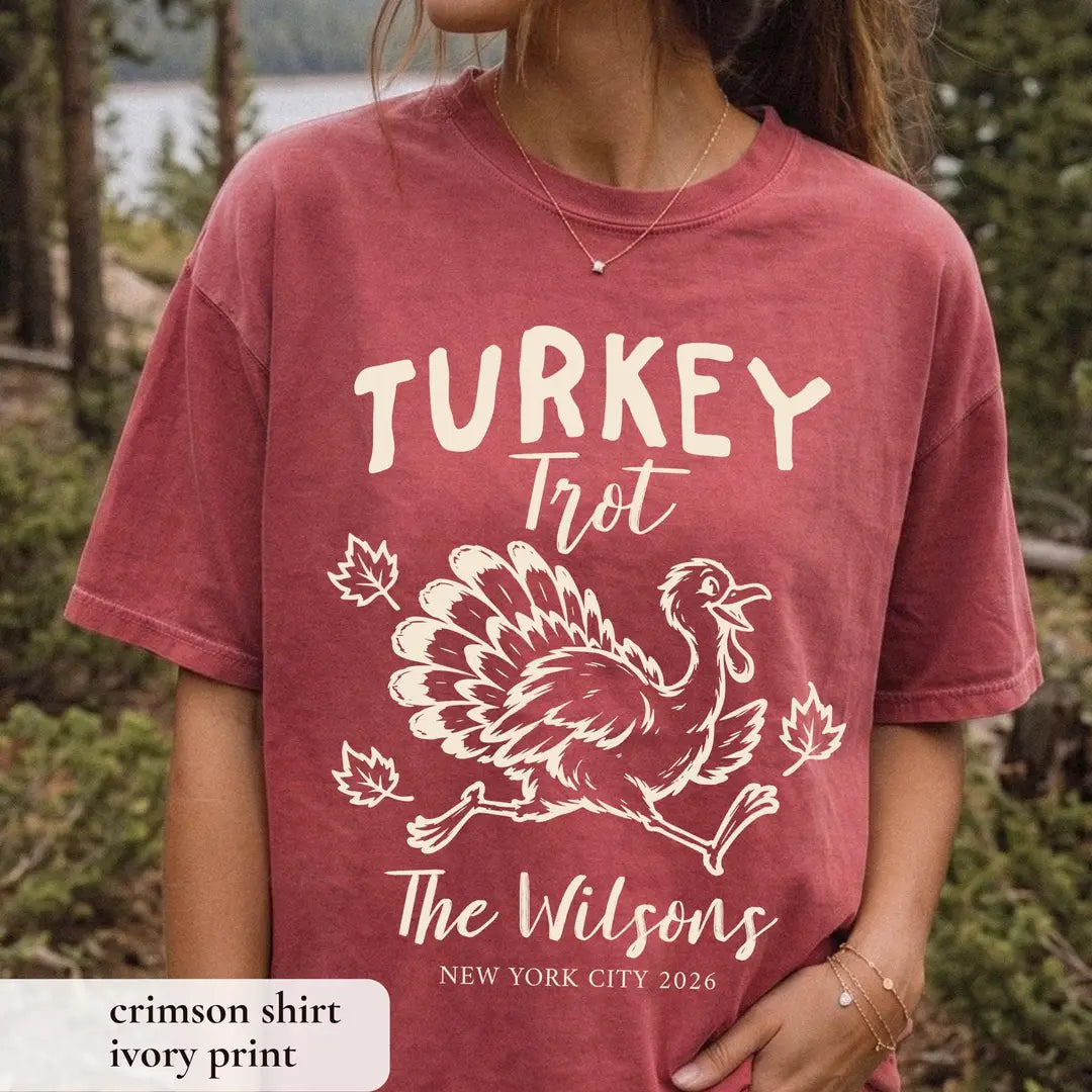 Woman in a Crimson Comfort Colors tee among pines by a lake, cream Turkey Trot turkey, leaves and New York City 2026