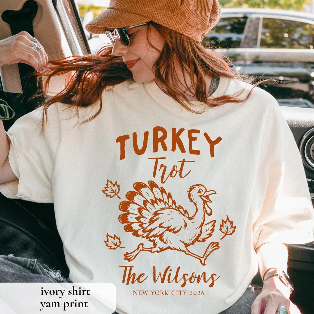 Close view of an Ivory Comfort Colors tee with rust Turkey Trot lettering, a running turkey, leaves and New York City 2026