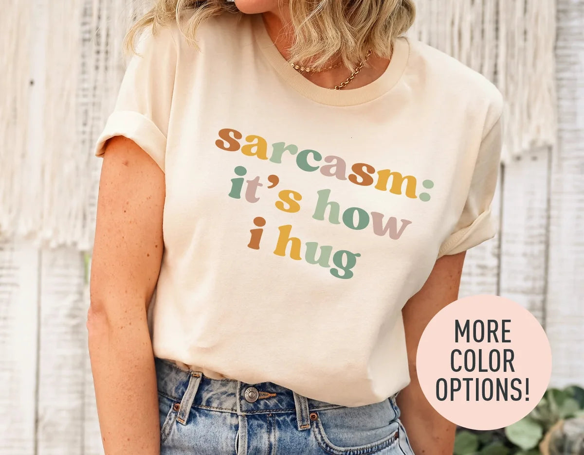 Woman in a natural cream tee with rolled sleeves and jeans, sarcasm: it's how i hug in sage, rust and rose retro type