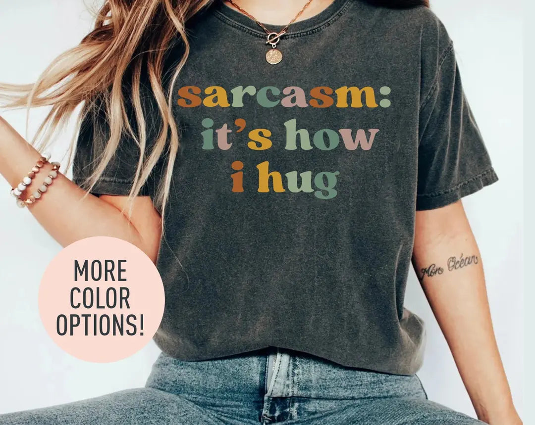 Woman in a dark gray Comfort Colors tee and jeans, bracelets on her wrist, with sarcasm: it's how i hug in retro letters