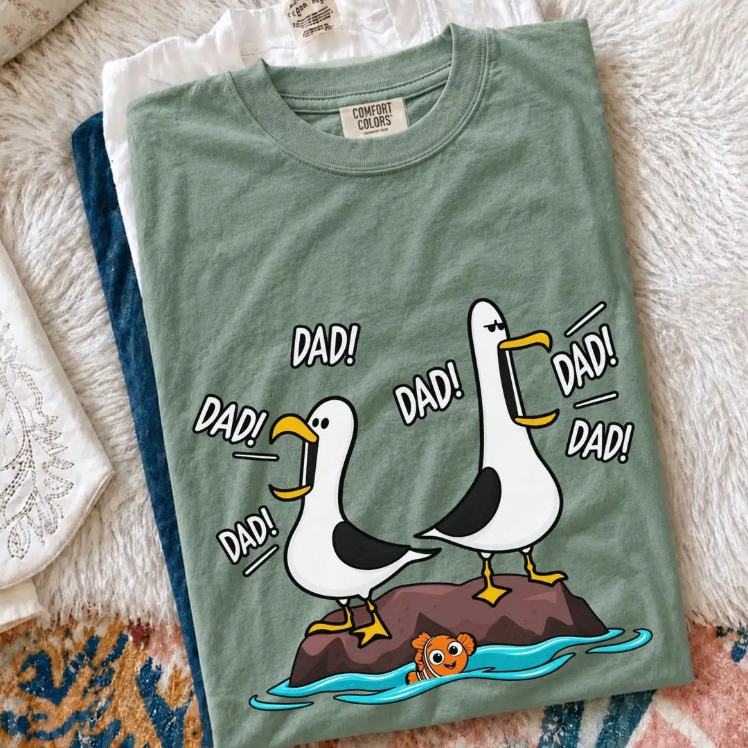 Folded Bay green Comfort Colors tee with two seagulls shouting Dad on a rock, laid on a shaggy rug with denim