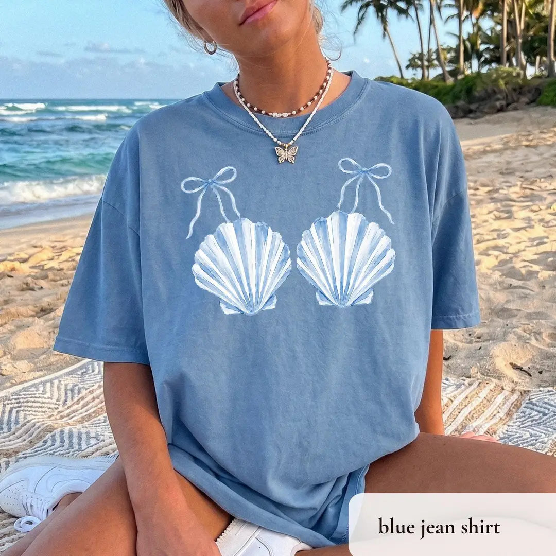 Woman in a Blue Jean Comfort Colors tee seated on a beach blanket, white striped scallop shells with tiny bows