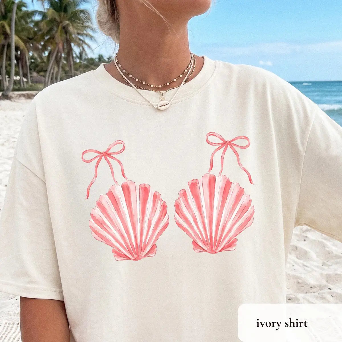 Ivory Comfort Colors tee with a beaded shell necklace, coral scallop shells and thin bow ties, palm trees behind