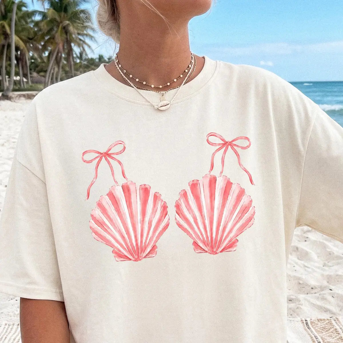 Woman in an Ivory Comfort Colors tee at the shore, coral striped scallop shells with tiny bows, pearl necklace