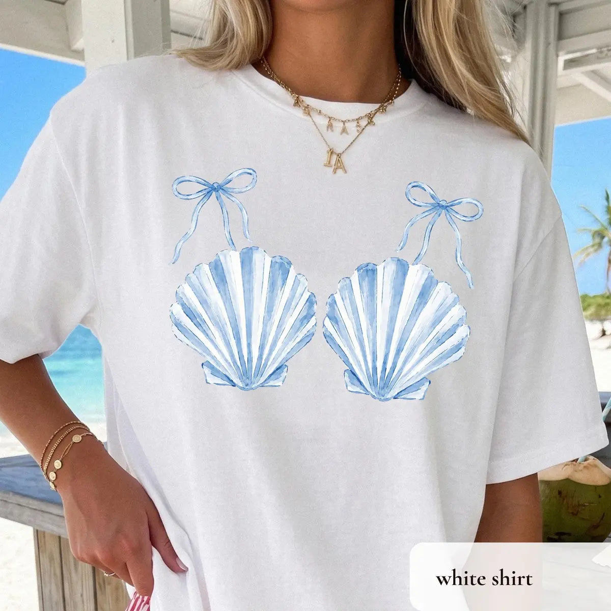 Woman in a White Comfort Colors tee on a beach porch, blue striped scallop shells with bows, gold bracelets