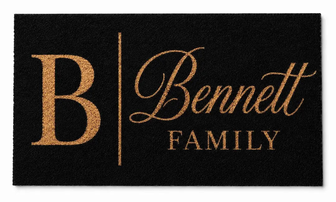 Flat view of a black faux coir doormat with a large gold-tone initial, script surname, and Family line