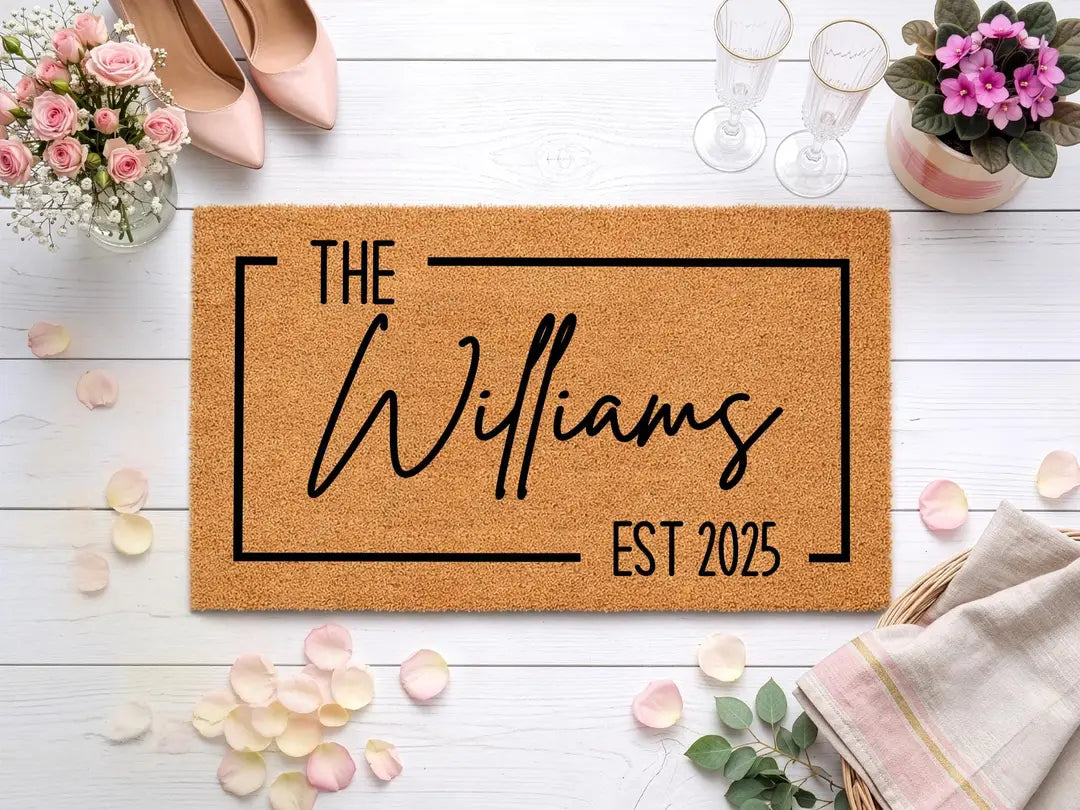 Rectangular coir doormat with a framed family name and established year styled with pink heels, champagne flutes, and rose petals