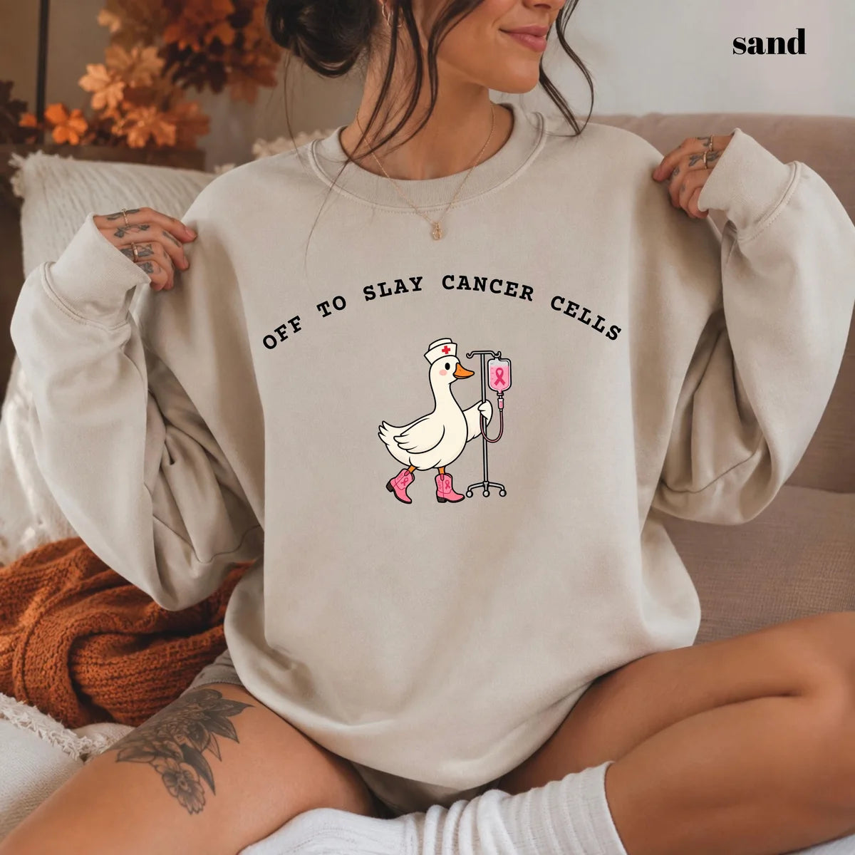 Sand crewneck sweatshirt with printed a nurse goose in pink boots with an IV pole and the words off to slay cancer cells, worn by a model.