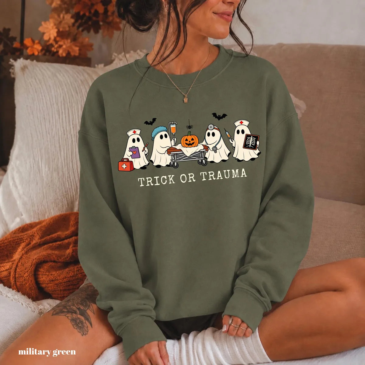 Military Green crewneck sweatshirt with printed nurse ghosts with a pumpkin and the words trick or trauma, worn by a model.