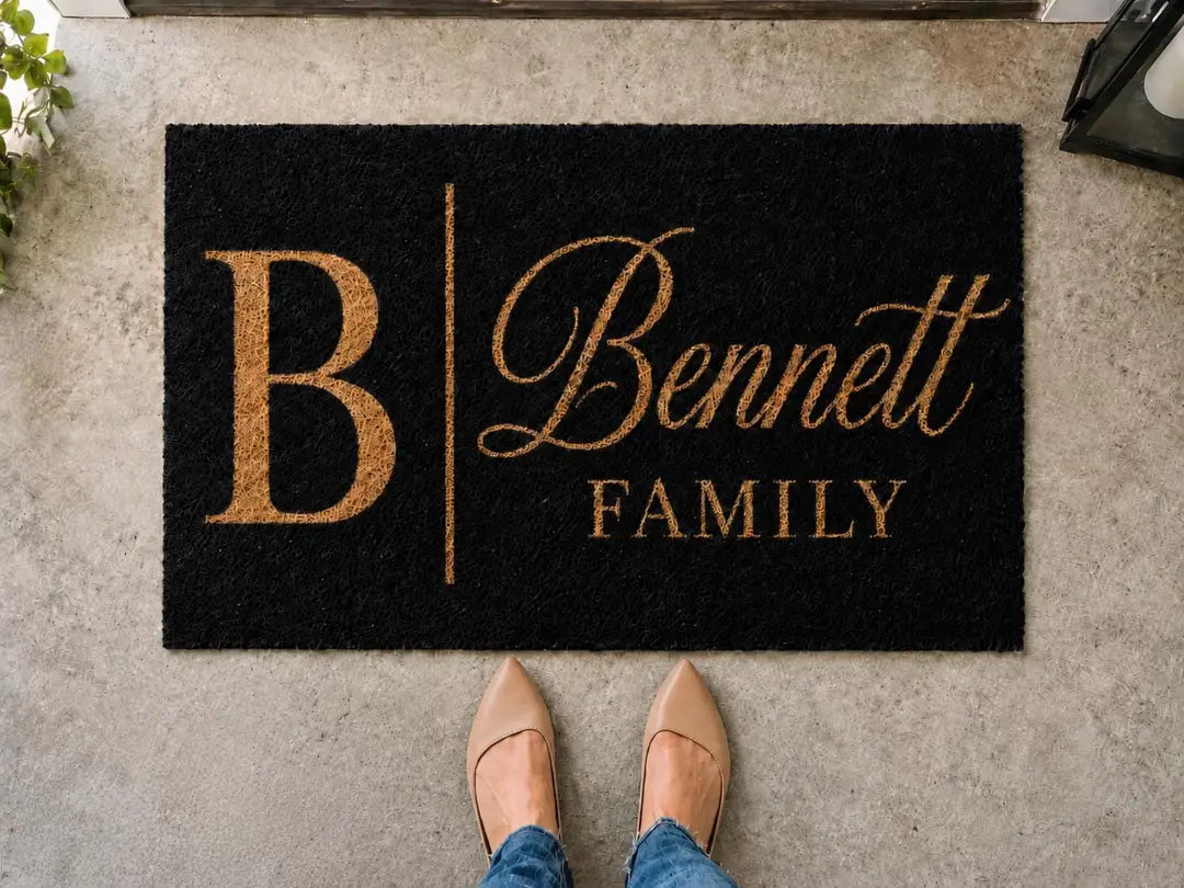 Black faux coir doormat with a gold-tone monogram initial, script surname, and Family line on a concrete step