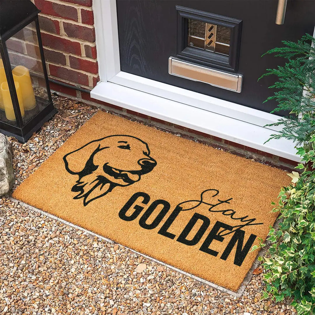 Rectangular coir doormat with a line-art golden retriever head and Stay Golden lettering on gravel in front of a black door