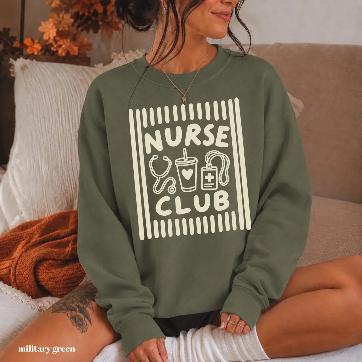 Military Green crewneck sweatshirt with printed a retro striped frame with a stethoscope, coffee cup and badge above the words nurse club, worn by a model.