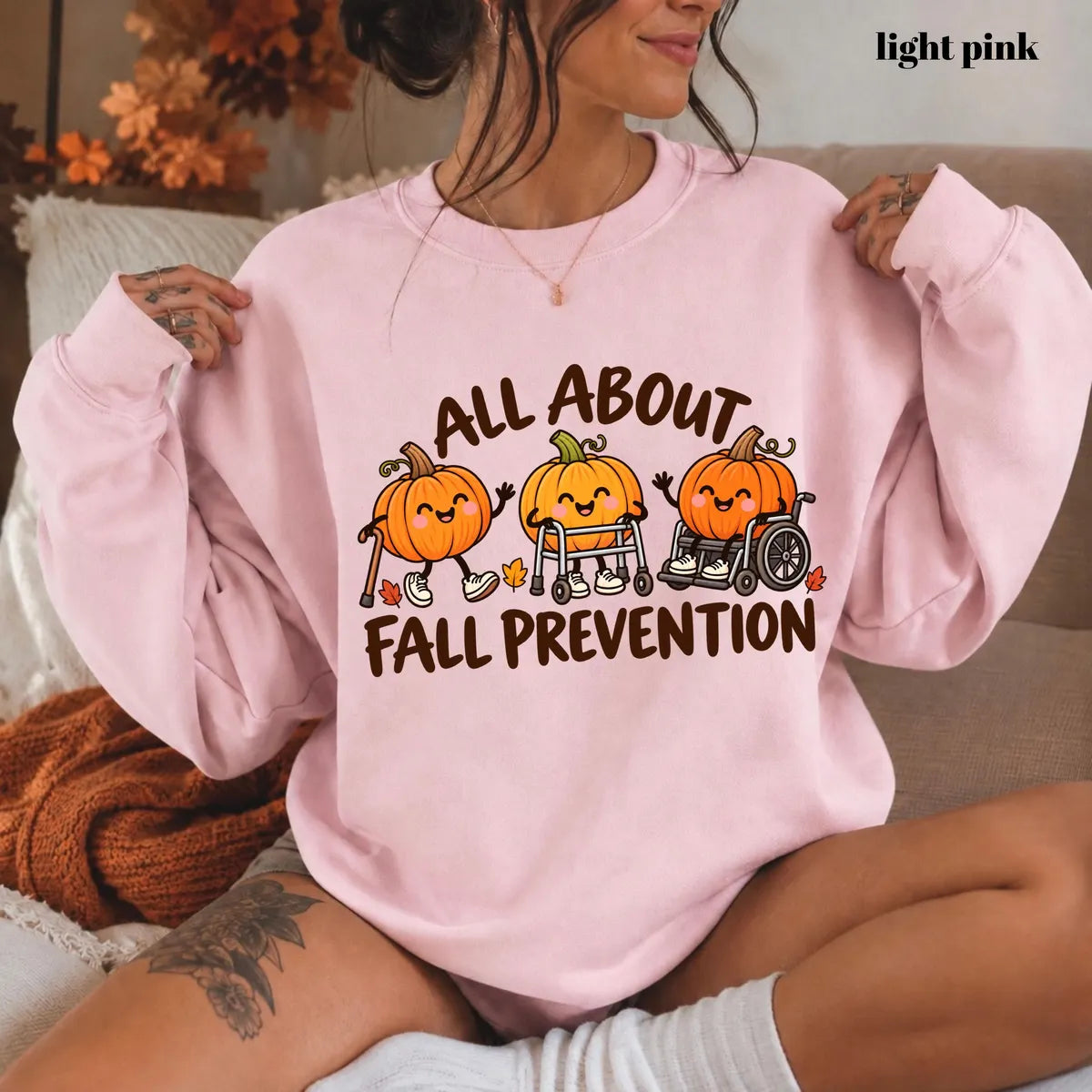 Light Pink crewneck sweatshirt with printed pumpkins with a walker and wheelchair above the words all about fall prevention, worn by a model.