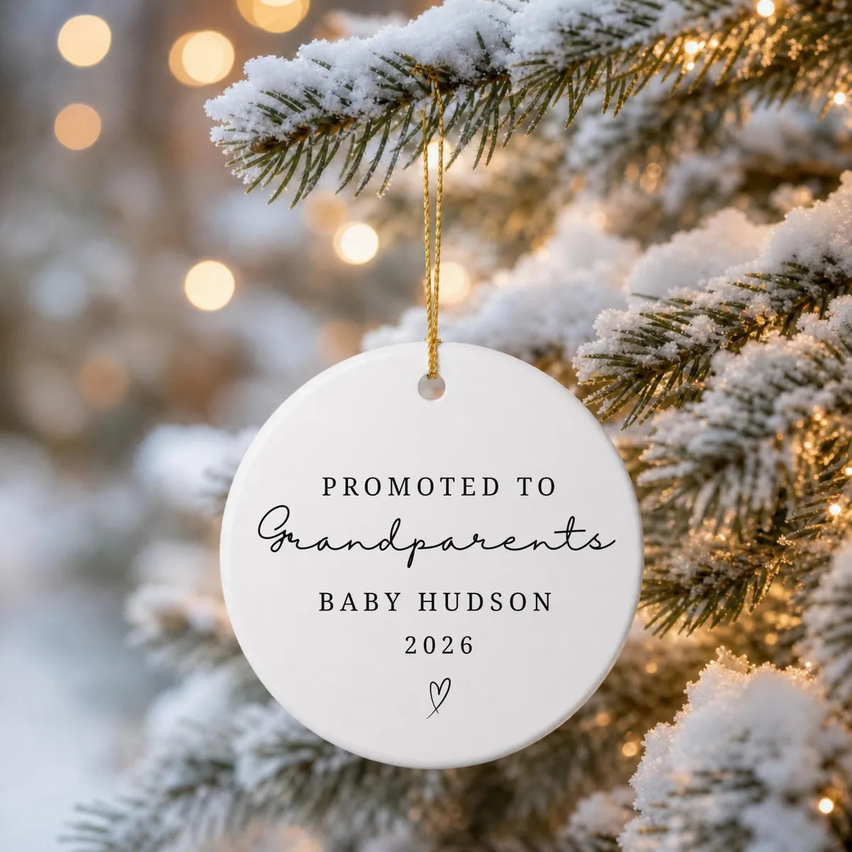 Round white ceramic Promoted to Grandparents ornament hanging from a snow-covered evergreen branch with warm string lights