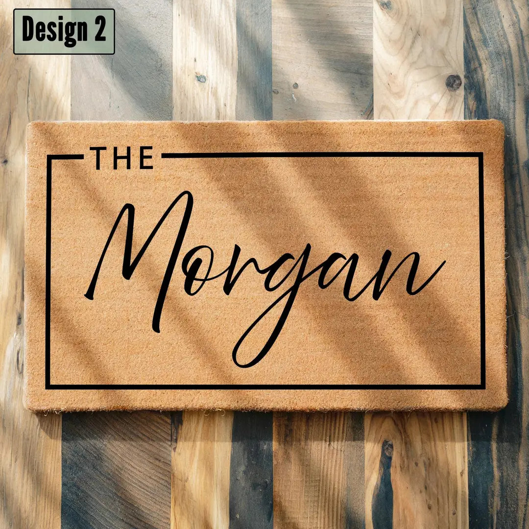 Rectangular coir doormat with a framed script family name on a mixed wood plank floor in dappled sunlight