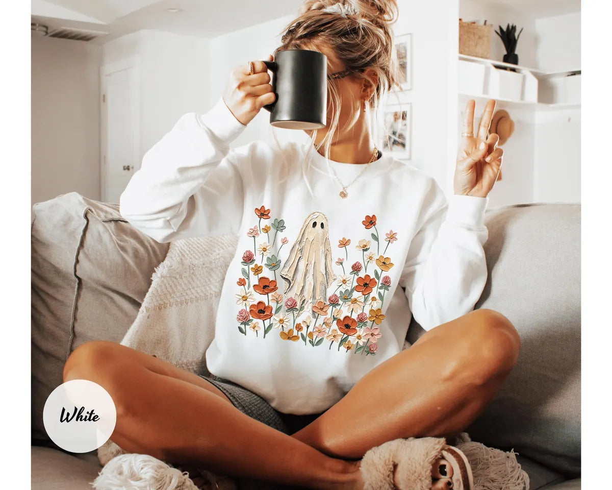White crewneck sweatshirt with printed a ghost standing in a field of autumn wildflowers, worn by a model.