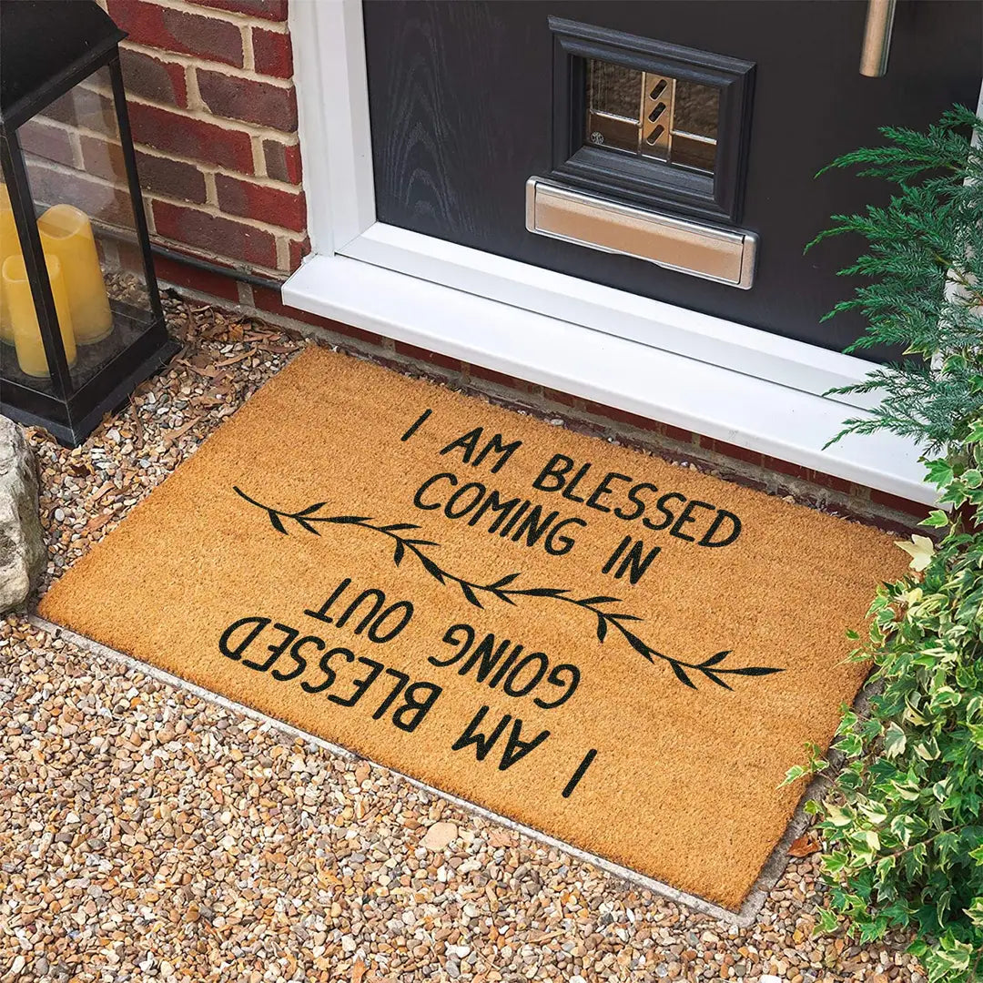 Rectangular coir doormat with I Am Blessed Coming In and Going Out lettering and a leaf branch on gravel in front of a black door