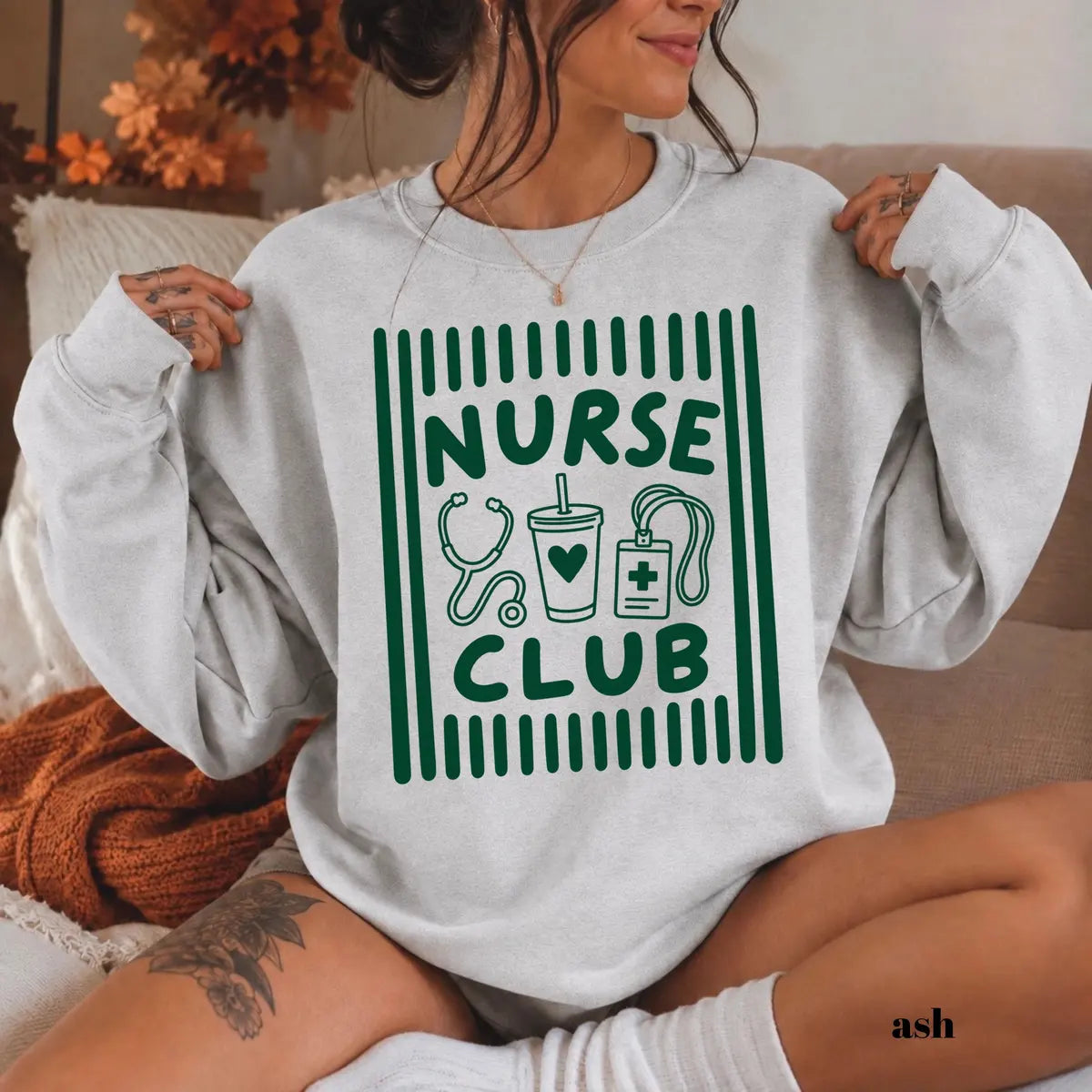 Ash crewneck sweatshirt with printed a retro striped frame with a stethoscope, coffee cup and badge above the words nurse club, worn by a model.