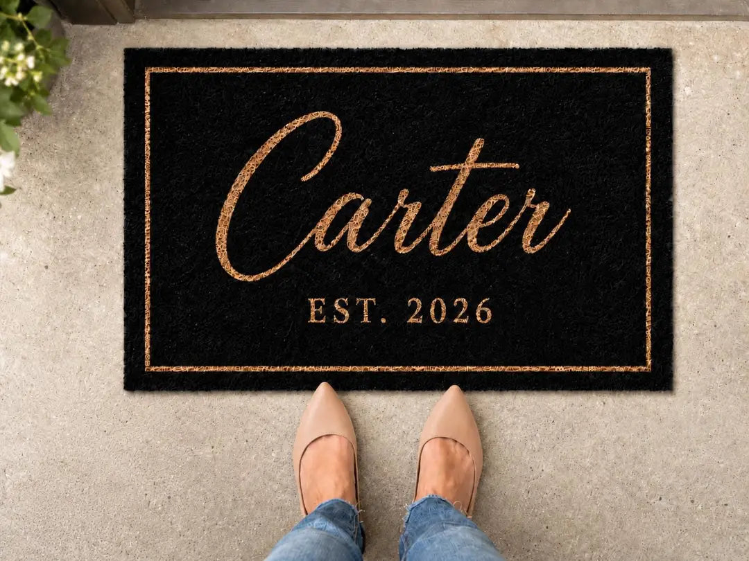 Black faux coir doormat with a gold-tone script surname, established year, and thin border on a concrete step