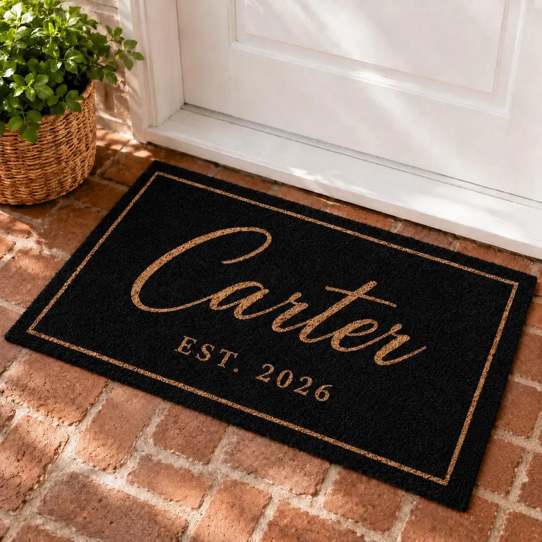 Rectangular black faux coir doormat with a gold-tone script surname, established year, and thin border at a white door on brick