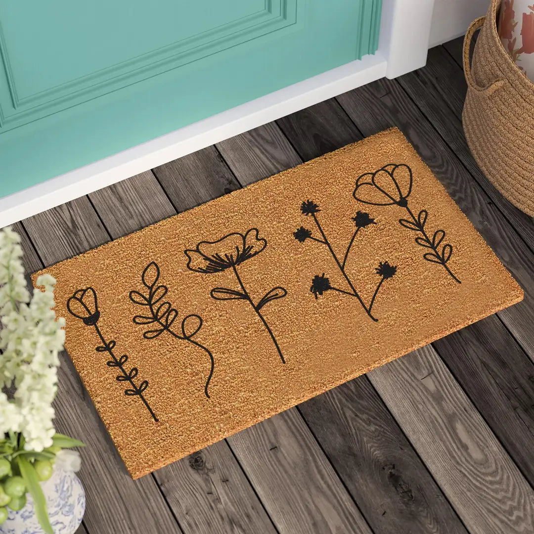 Coir doormat with black line-art wildflowers and leafy stems on a wooden porch beside a teal door and woven basket
