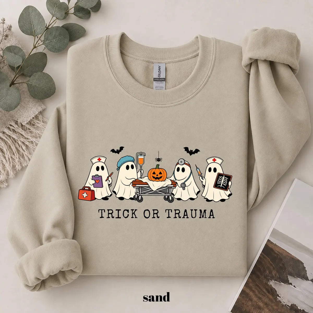 Sand crewneck sweatshirt with printed nurse ghosts with a pumpkin and the words trick or trauma, flat lay view.