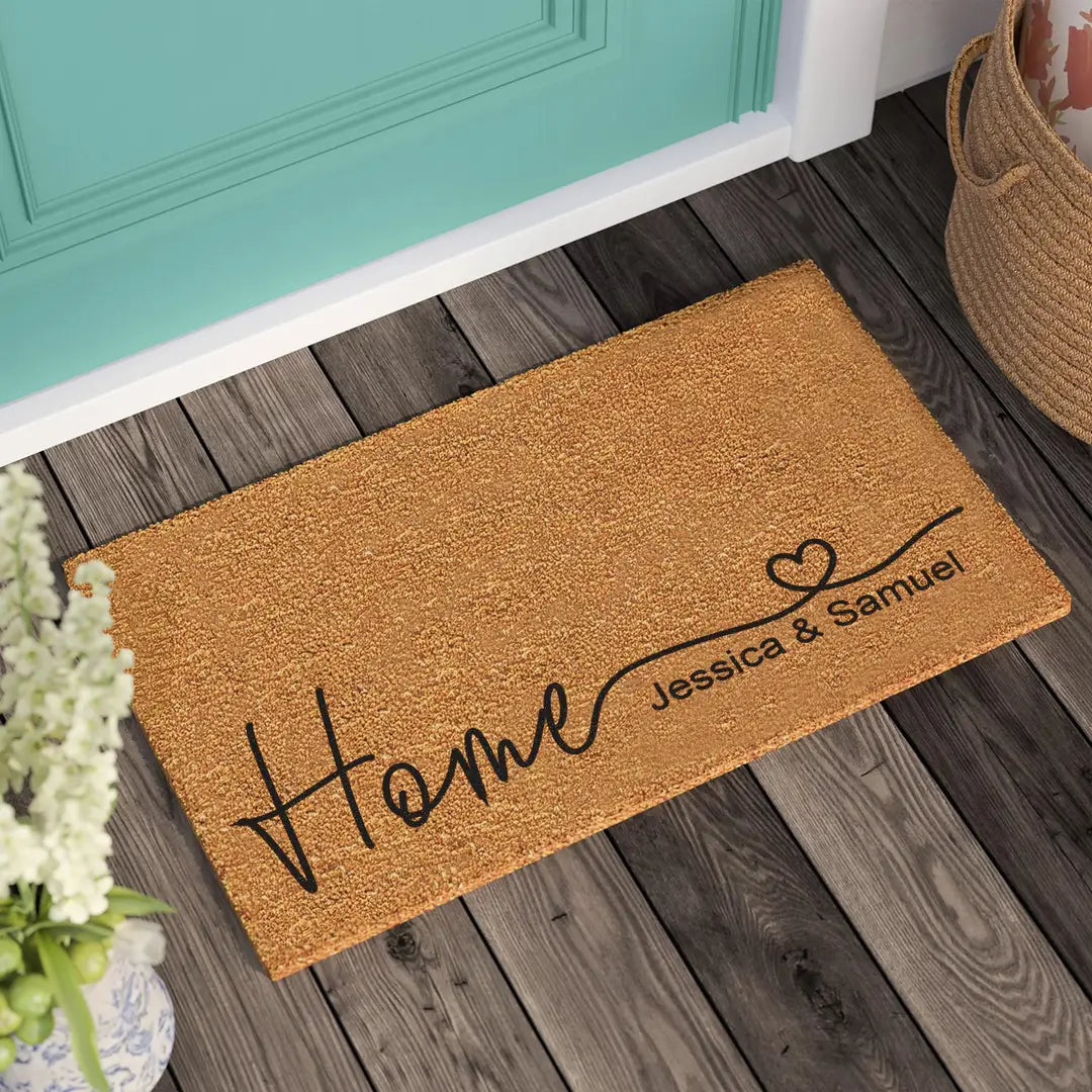 Coir doormat with a heart script Home and two names on a wooden porch beside a teal door and woven basket