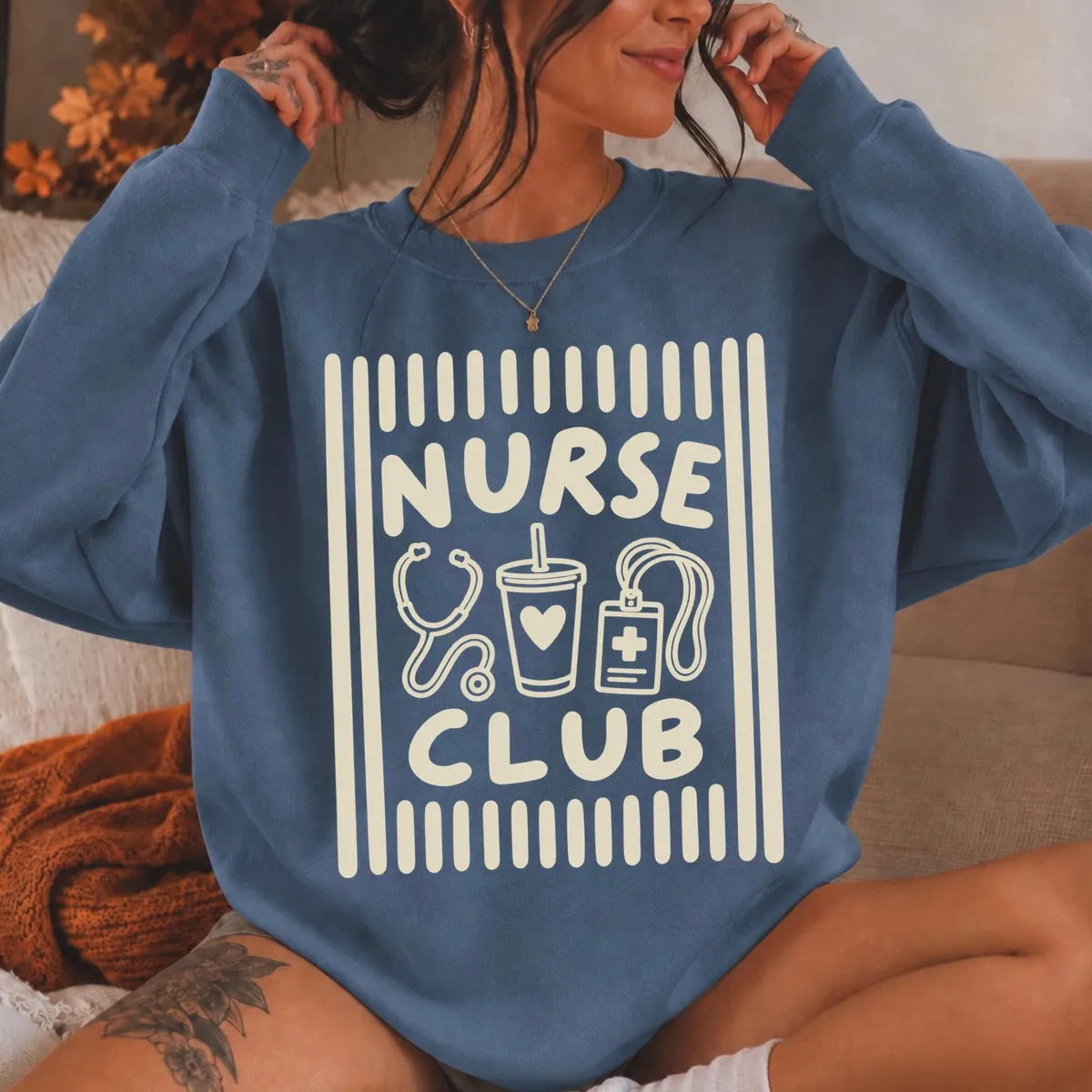 Indigo Blue crewneck sweatshirt with printed a retro striped frame with a stethoscope, coffee cup and badge above the words nurse club, worn by a model.