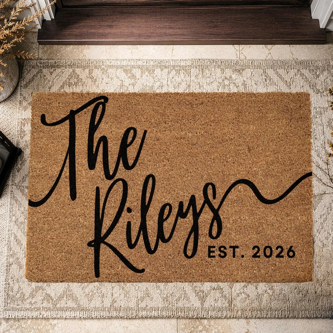 Coir doormat with a script family name and established year layered on a beige patterned rug beside a doorstep and dried grass
