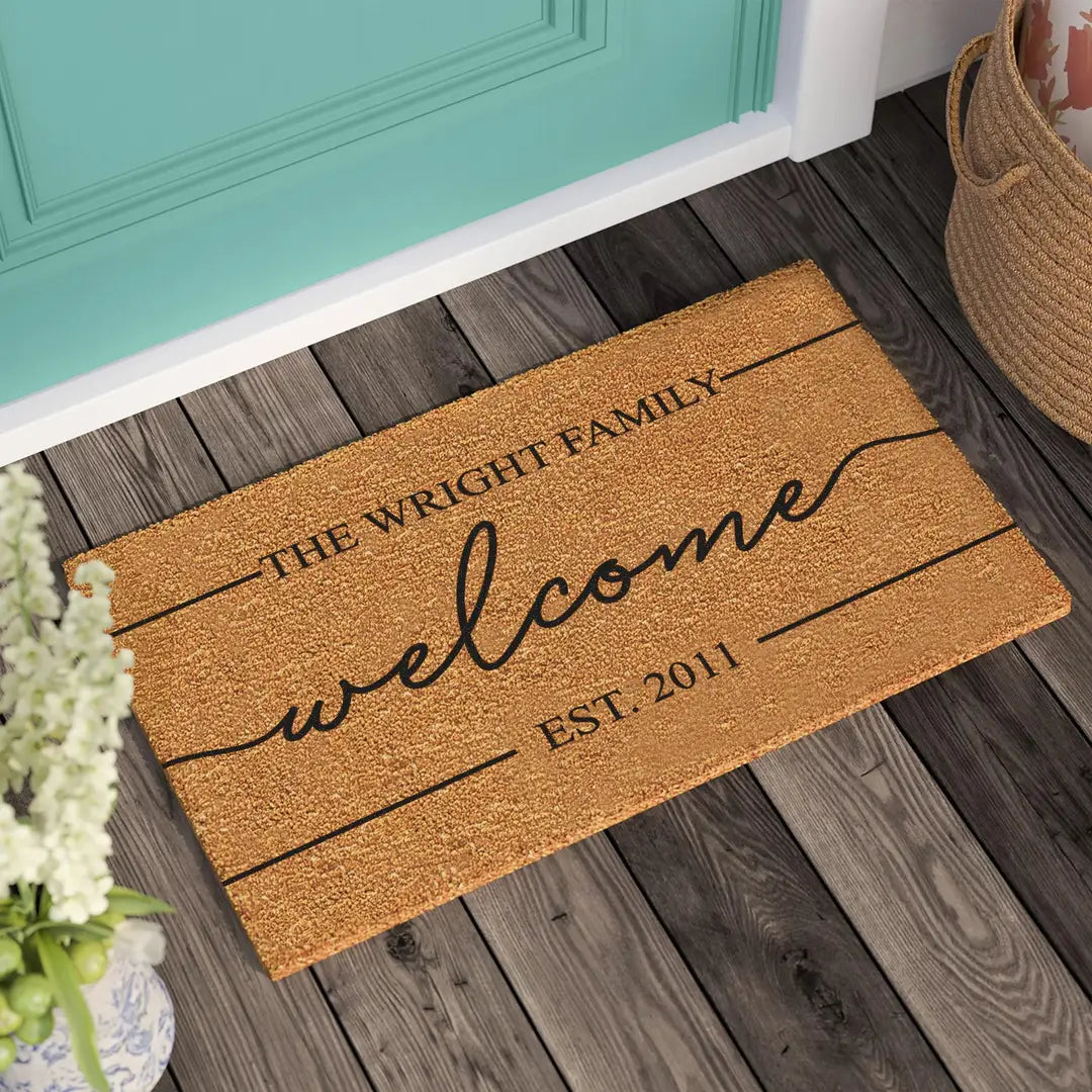 Coir doormat with a modern script welcome, family name, and established year on a wooden porch beside a teal door and woven basket