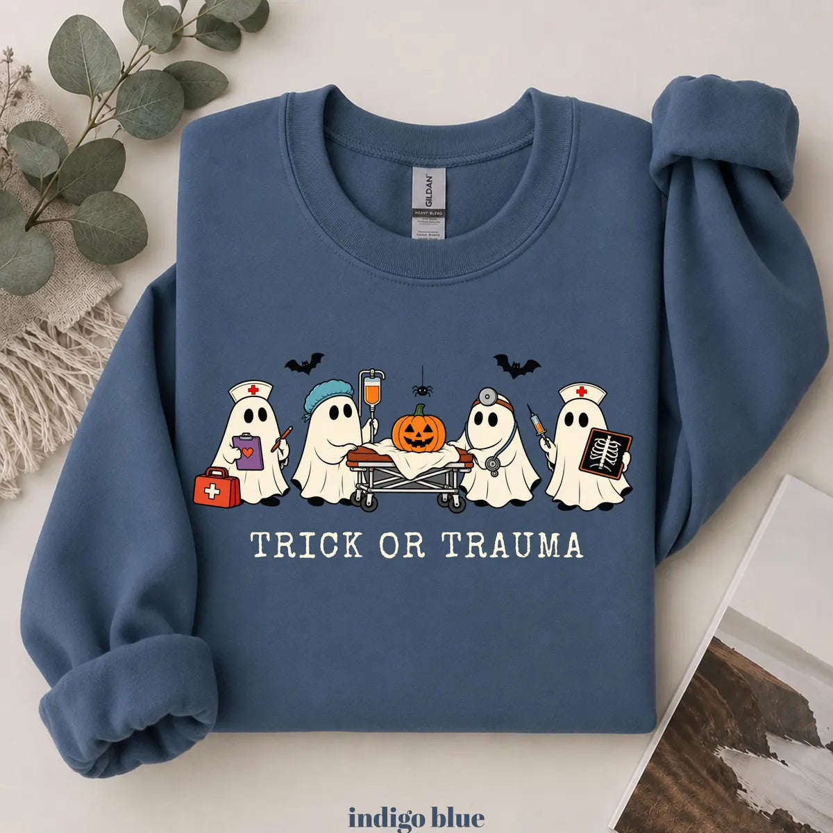 Indigo Blue crewneck sweatshirt with printed nurse ghosts with a pumpkin and the words trick or trauma, flat lay view.