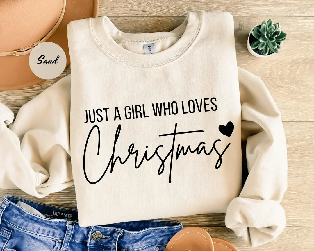 Sand crewneck sweatshirt with script lettering that reads just a girl who loves Christmas with a small heart, flat lay view.