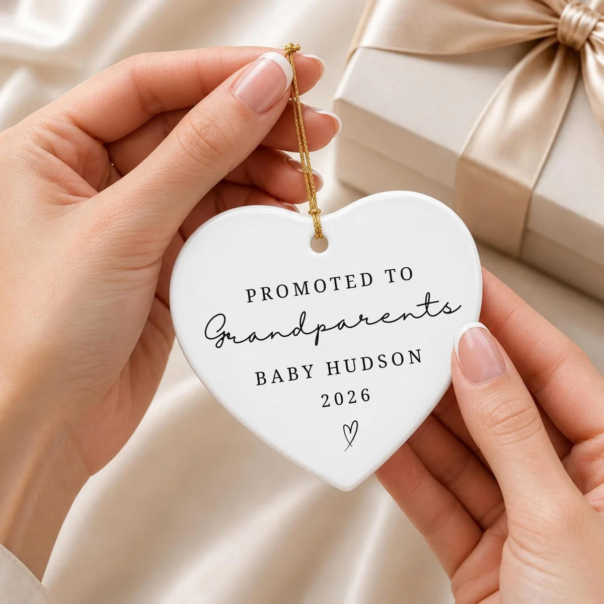 Hands holding a heart-shaped white ceramic Promoted to Grandparents ornament by its gold string beside a wrapped gift