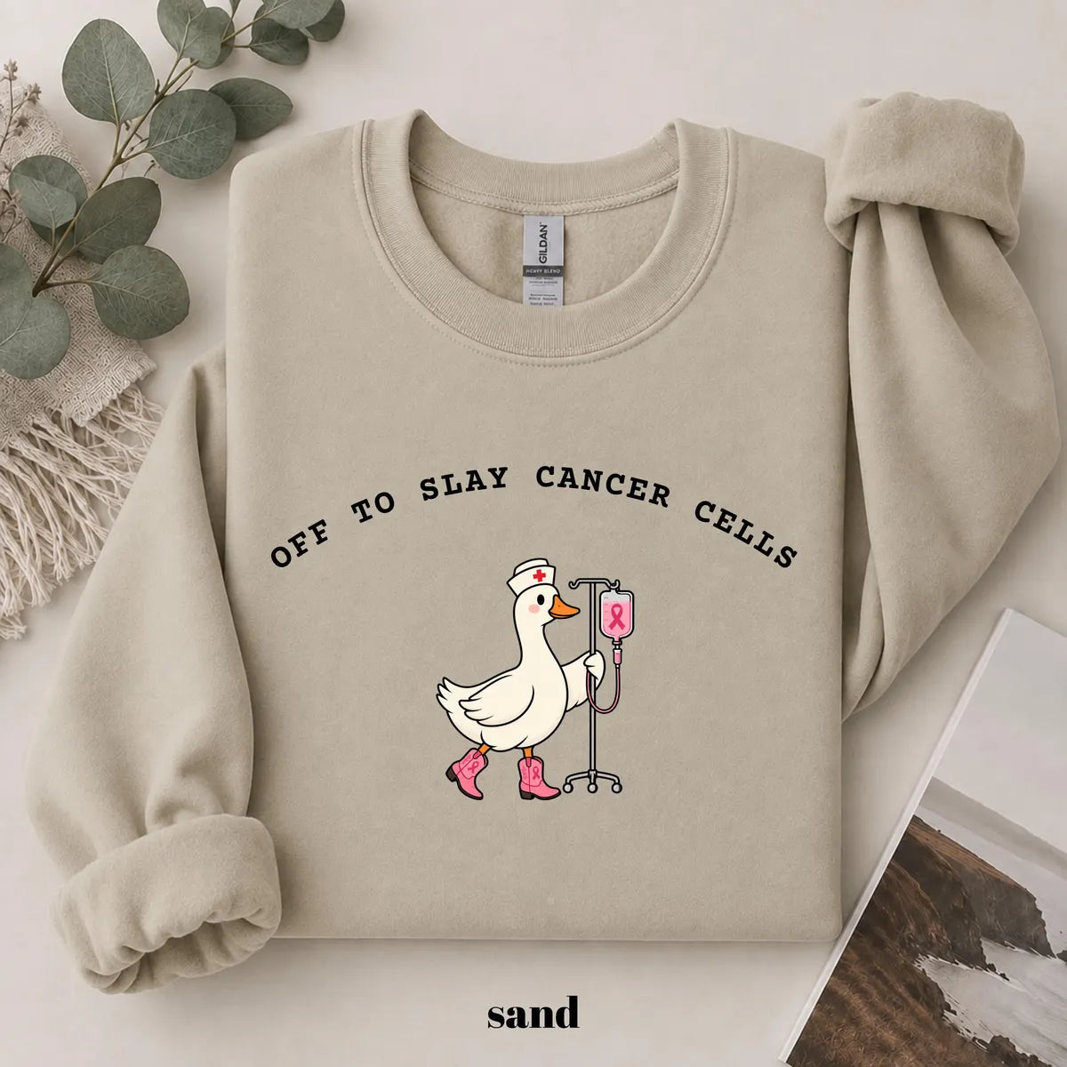 Sand crewneck sweatshirt with printed a nurse goose in pink boots with an IV pole and the words off to slay cancer cells, flat lay view.