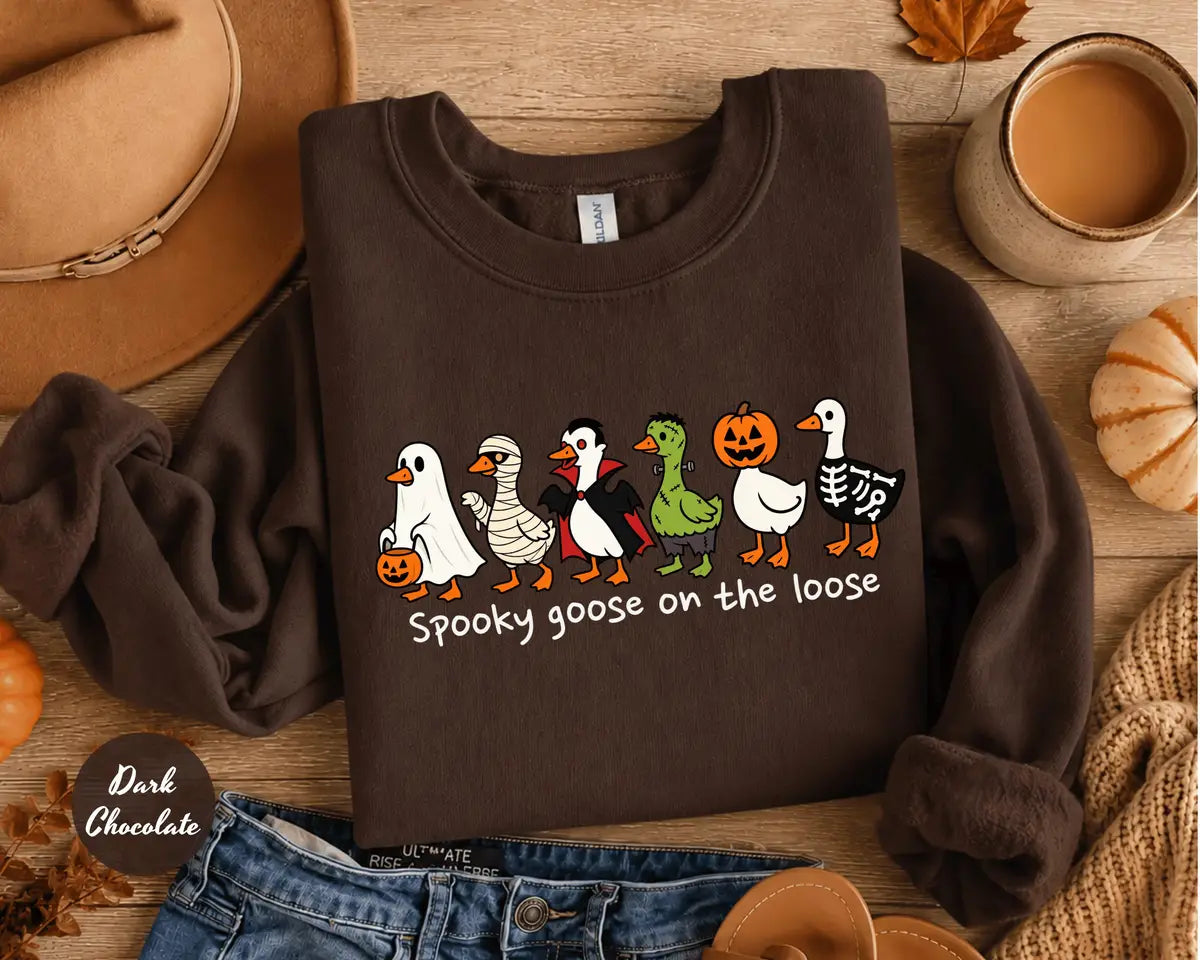 Dark Chocolate crewneck sweatshirt with costumed geese dressed as a ghost, vampire, pumpkin and skeleton above the words spooky goose on the loose, flat lay view.