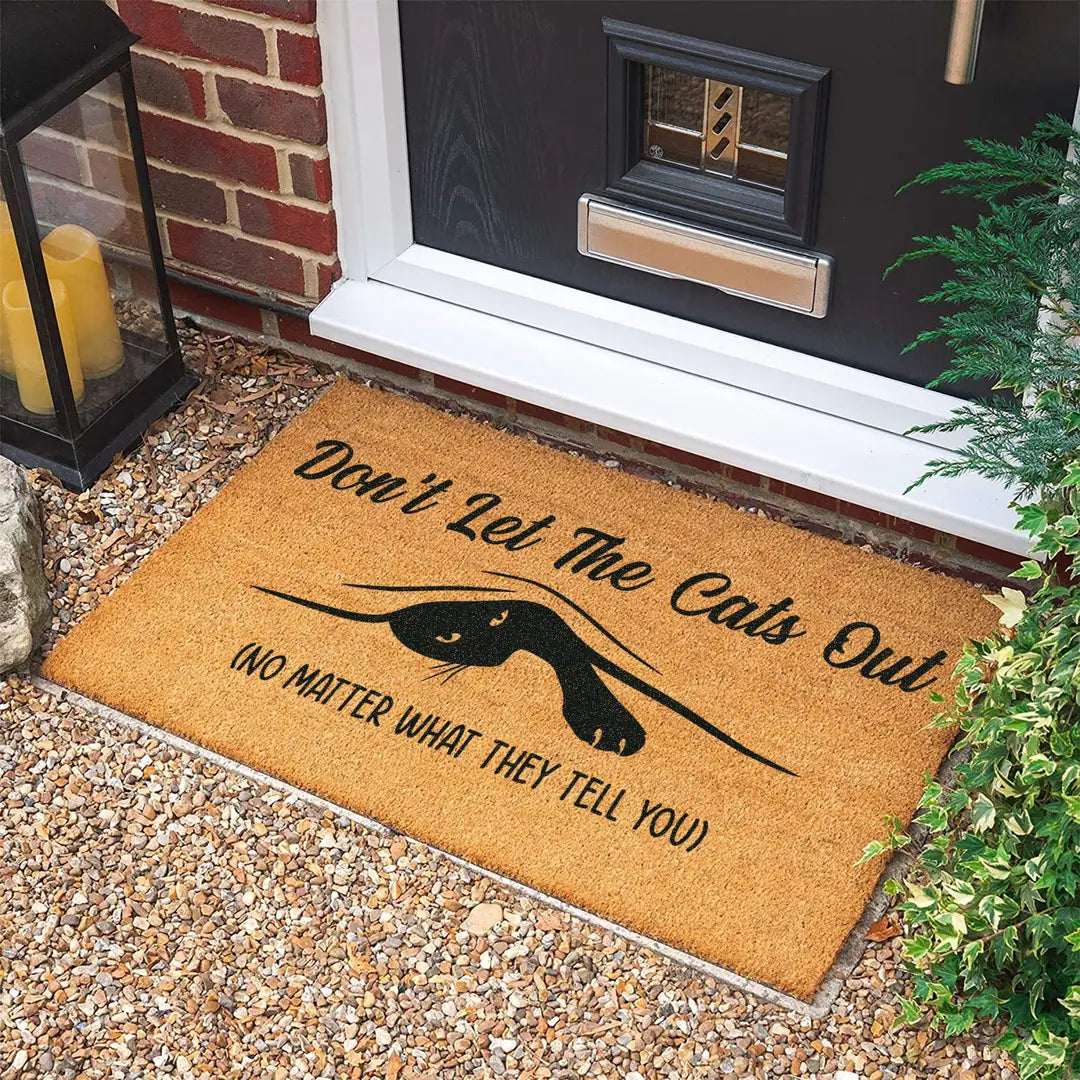 Coir doormat with a peeking black cat and the message Don't Let The Cats Out at a dark front door on gravel