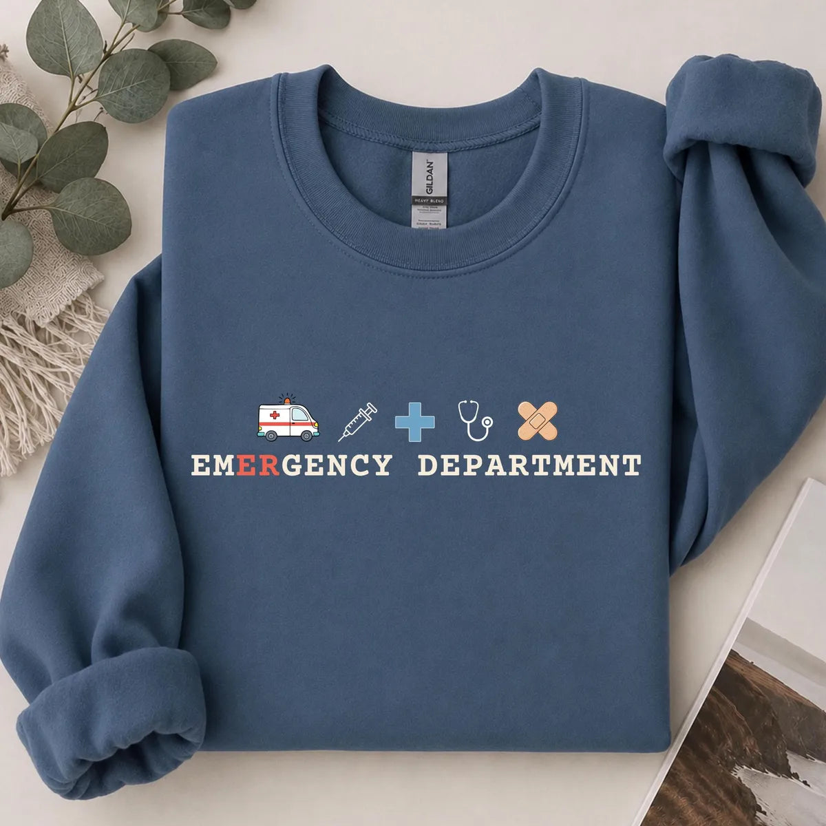 Indigo Blue crewneck sweatshirt with printed an ambulance, syringe, cross, stethoscope and bandage above the words emergency department, flat lay view.