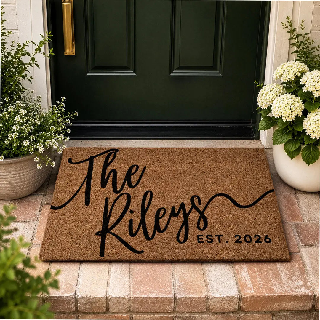 Rectangular coir doormat with a script family name and established year at a dark green door on brick steps with white hydrangeas
