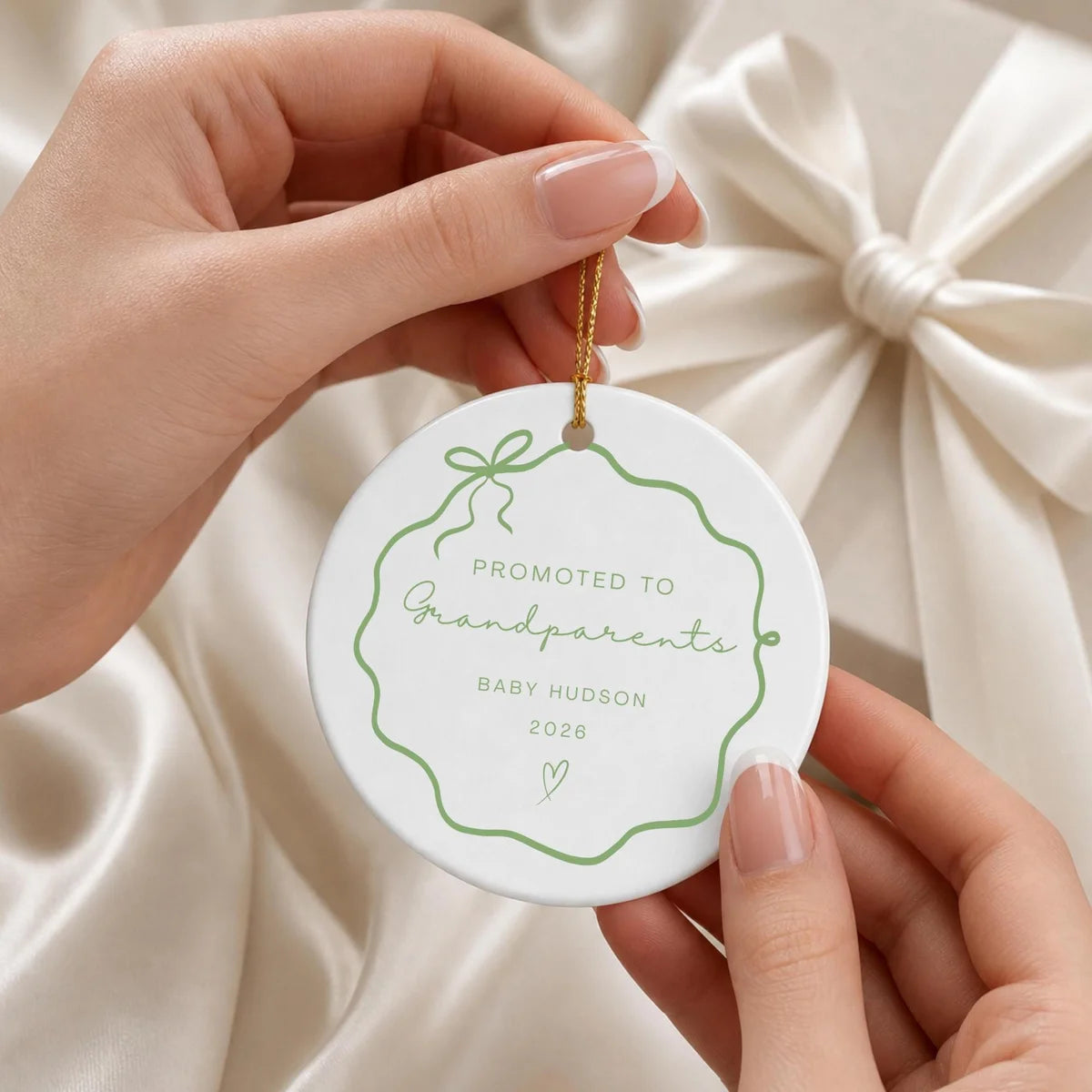 Hands holding a green wavy bow frame round ceramic Promoted to Grandparents ornament by its gold string in front of a cream gift bow
