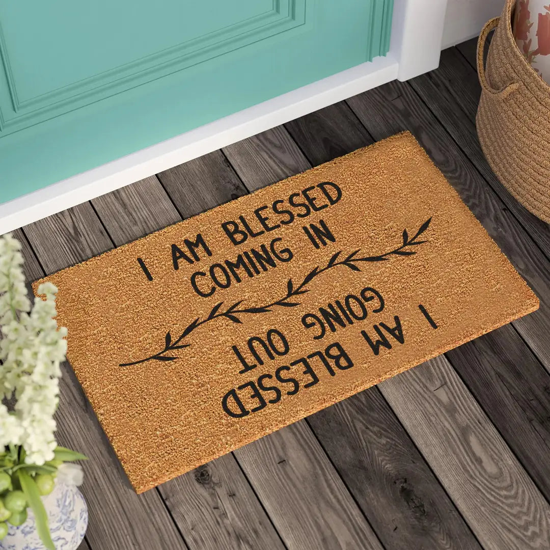 Coir doormat with I Am Blessed Coming In and Going Out lettering and a leaf branch on a wooden porch beside a teal door