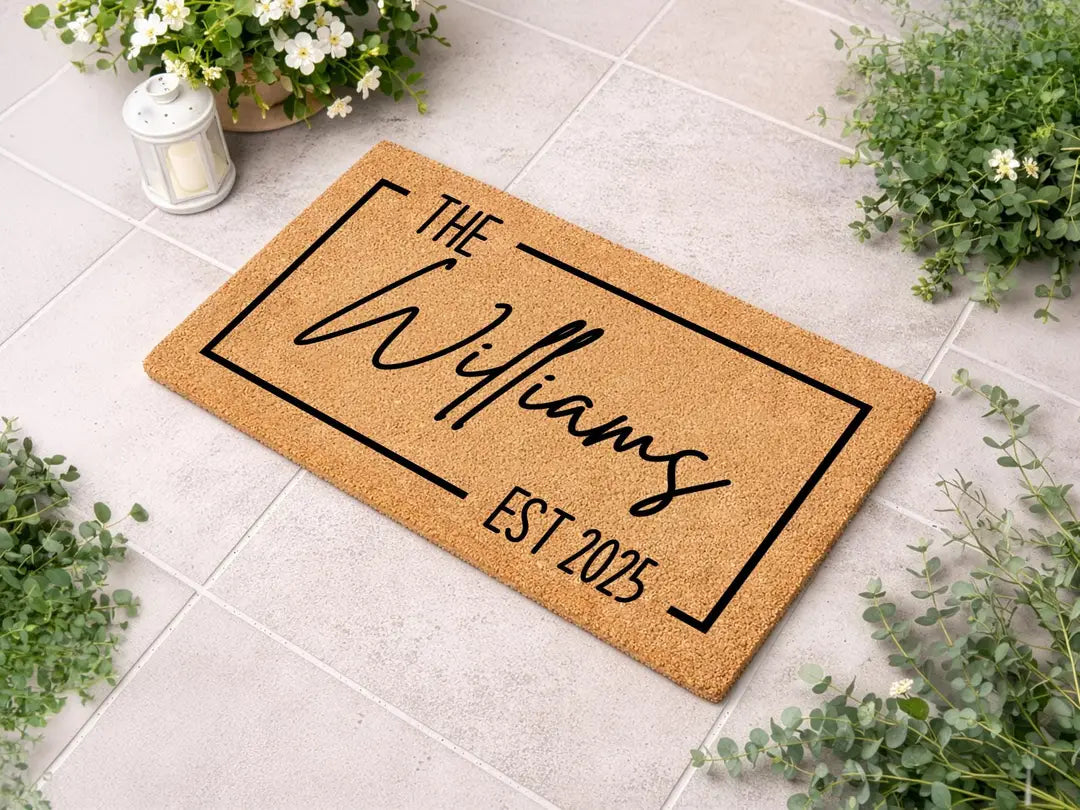 Coir doormat with a framed family name and established year on pale tile with greenery and a lantern