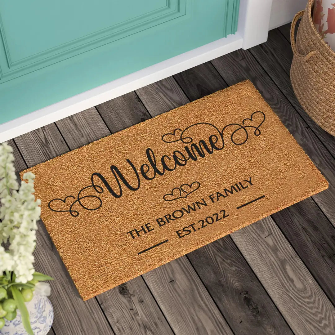 Coir doormat with a heart script Welcome, family name, and established year on a wooden porch beside a teal door and woven basket