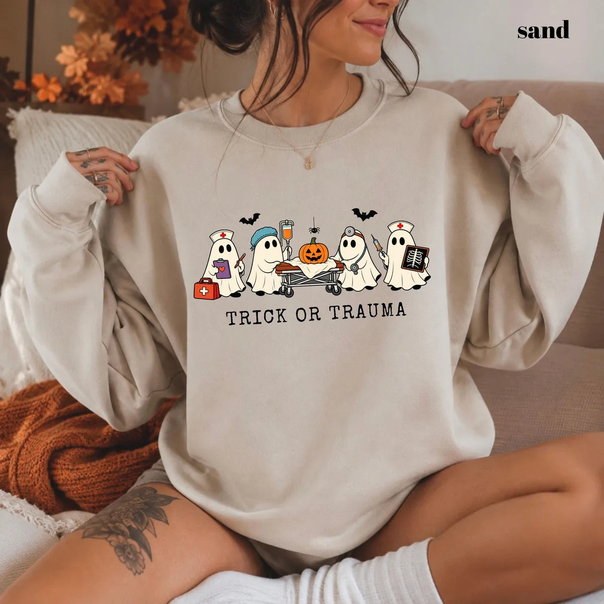 Sand crewneck sweatshirt with printed nurse ghosts with a pumpkin and the words trick or trauma, worn by a model.